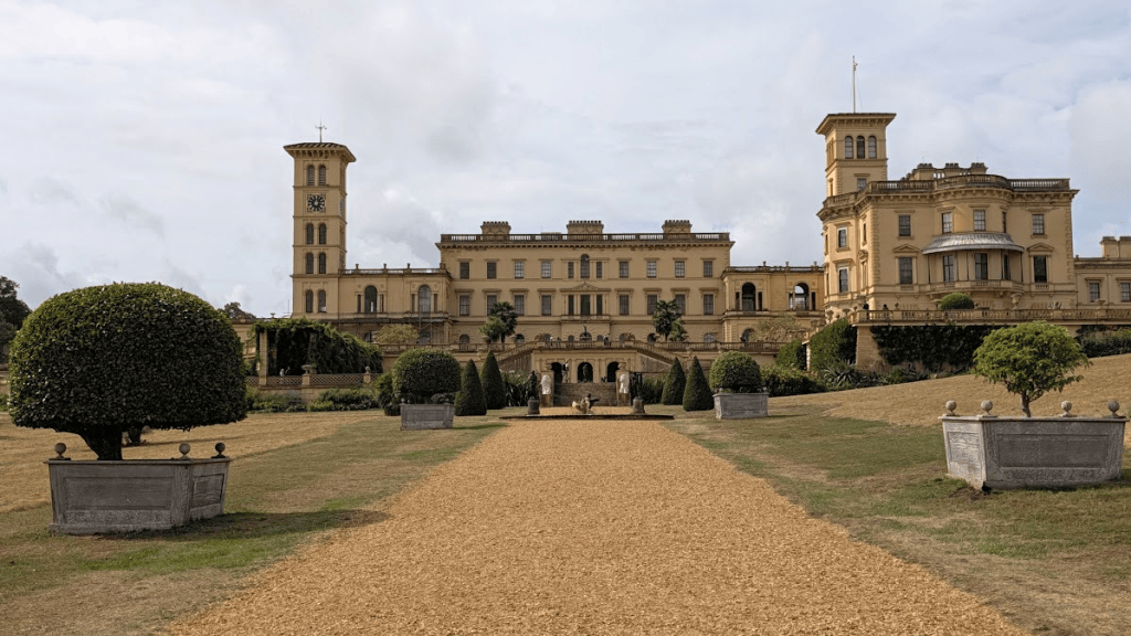 A grand historical building with a yellow facade, featuring multiple towers and a clock, surrounded by neatly trimmed gardens and topiary. A gravel pathway leads to the entrance.