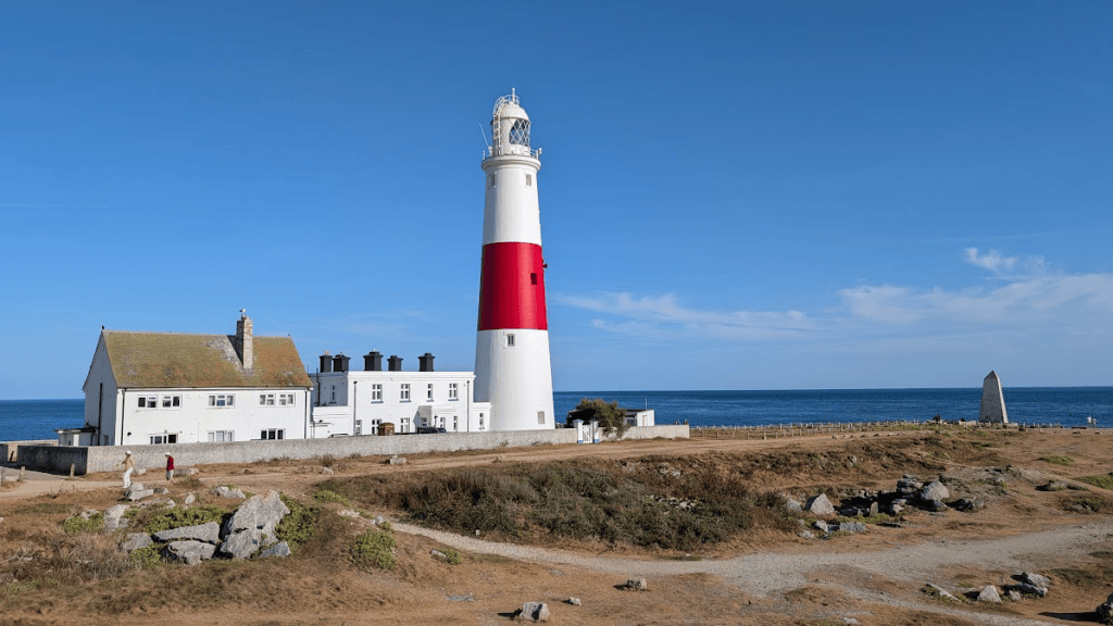 A coastal lighthouse with a white and red striped tower next to a white house, set against a clear blue sky and ocean.