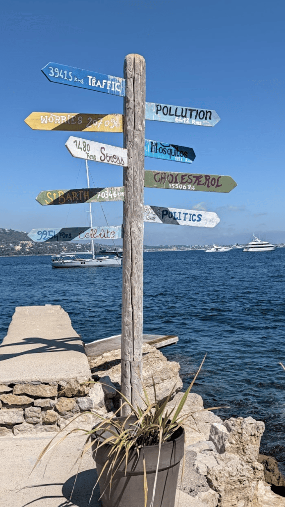 A wooden signpost with colorful arrows pointing in different directions, each labeled with various words and distances like 'Traffic', 'Pollution', ' stress', and 'Politics', situated by the waterfront with a view of boats and clear blue skies.