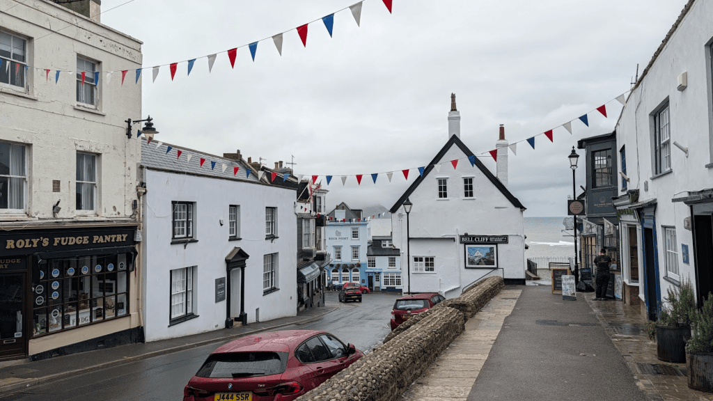 A seaside street view featuring white buildings with bunting, a shop named Roly's Fudge Pantry, and a glimpse of the ocean in the background.