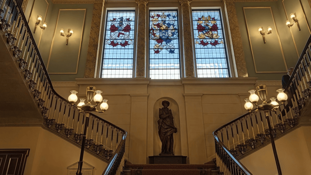 Interior view of a grand staircase featuring stained glass windows, ornate railings, and a statue at the base of the stairs.